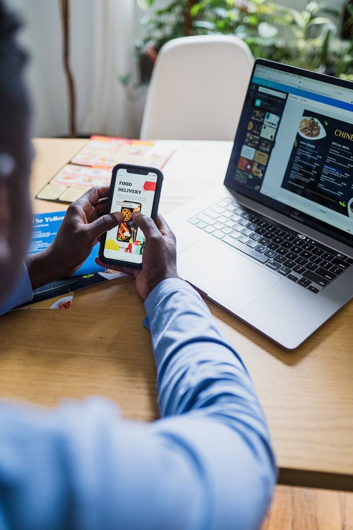 A man uses a smartphone and laptop to order food online, showcasing modern technology.